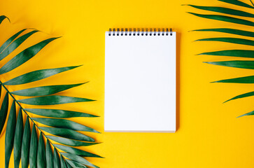 A blank spiral notebook on a yellow background with green palm leaves. Creating a tropical and minimalist design.