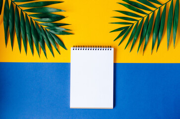 A blank spiral notebook on a yellow background with green palm leaves. Creating a tropical and minimalist design.