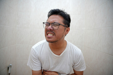 Obraz premium Asian man sitting on the toilet holding his stomach with a pained expression, symbolizing stomachache or discomfort, conveying human emotions.