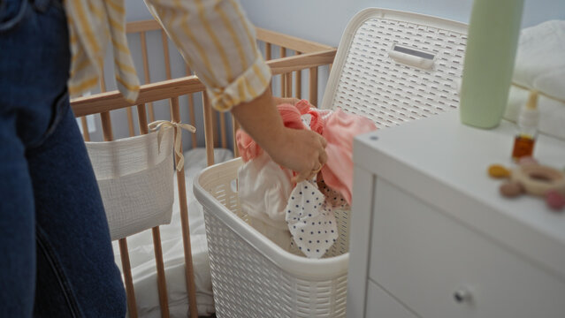 Woman handling laundry in a home room, showcasing clothing in a basket near a crib, highlighting indoor cleaning chores and personal care tasks for young infants.