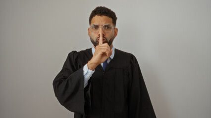Young man with glasses in graduation gown making shushing gesture over white background, emphasizing silence