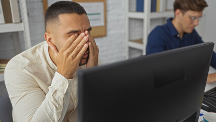 Frustrated man covering his face with hands while working on a computer in an office alongside...
