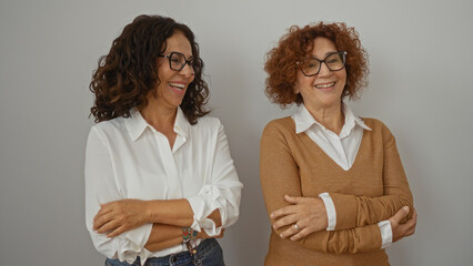 Two middle-aged hispanic women smiling warmly, standing together with arms crossed against a white...