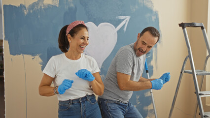 Middle-aged hispanic couple dances joyfully home interior while painting new house wall together, symbolizing love and family in the living room atmosphere.