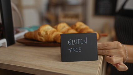 Woman holding sign in a bakery showcasing gluten-free croissants on a wooden counter with an indoor...