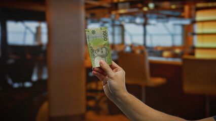 Hand holding a chilean peso banknote in a dimly lit indoor coffee shop, symbolizing business investment and monetary exchange in a relaxed setting.