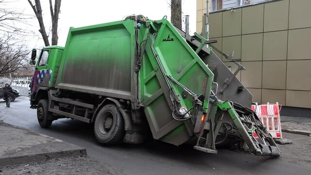 A green garbage truck lies on its side on a city street after falling into a hole. orange warning netting reveals a hole in the asphalt road. A hole in the road.