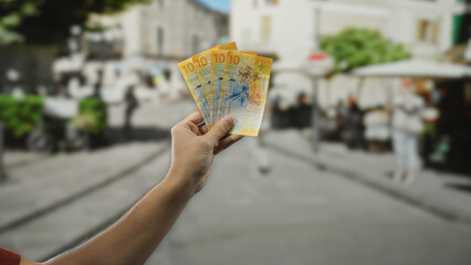 Man holding swiss franc banknotes on a city street with blurred pedestrians and buildings in the...