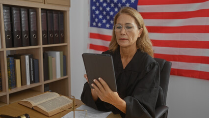 Woman judge in black robe using tablet with american flag background in office with books and gavel...