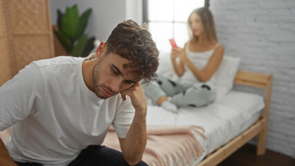 Man sitting on bed in bedroom while woman uses phone, showcasing home relationship dynamics and...