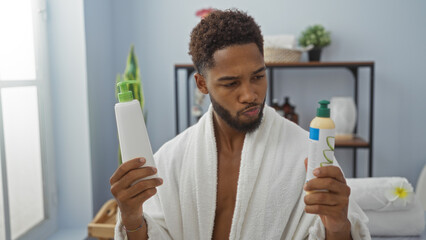 Young man in a spa wearing a bathrobe and holding two bottles of beauty products, contemplating...