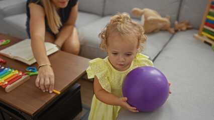 Fototapeta premium Woman supervising toddler girl holding purple ball in an indoor living room setting with toys and open book on table