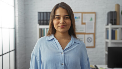Fototapeta premium Young hispanic woman smiling confidently in a modern office environment with shelves and pinned documents in the background