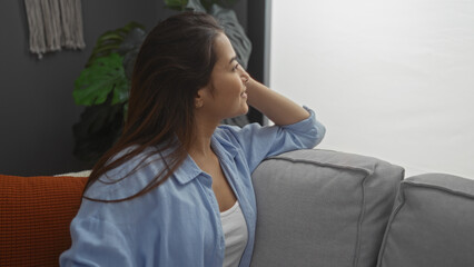 Beautiful young hispanic woman in a living room, sitting on a couch and looking out a bright window, indoor setting.