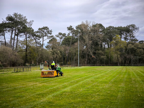 Operador no seu trator cortador de grama em amplo campo desportivo, cercado por &aacute;rvores altas sob um c&eacute;u nublado