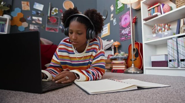 Ground level shot of concentrated Black gen z girl in headphones doing school assignment lying on floor in her room, working on laptop and studying online at home