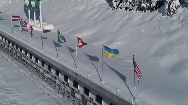 Flags representing various countries flutter in the cold winter breeze, surrounded by pristine snow. The scene highlights an Olympic location, showcasing international unity.