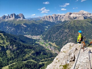 A climber in a bright helmet pauses atop a rugged cliff, gazing over the lush green valley and...