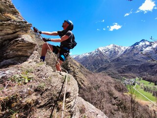 Via Ferrata di Rocca Candelera, Usseglio: A rock climber conquers a craggy peak, with snow-capped mountains rising majestically against a backdrop of bright blue sky.