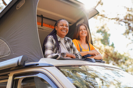 Multiracial elderly female friends enjoy day in nature with mini van camper - Happy mature women during road trip vacation in the woods