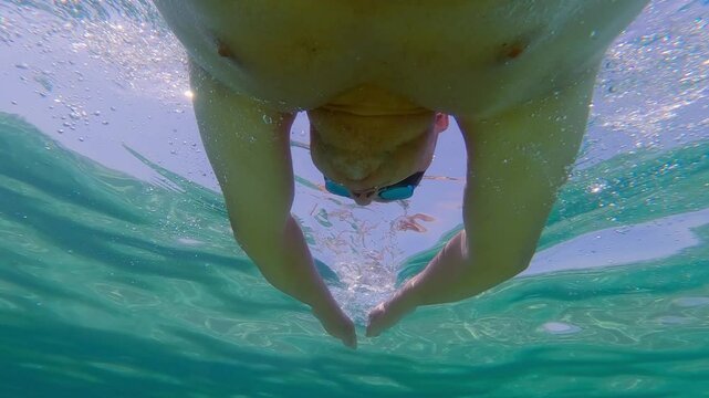 Underwater perspective of a man swimming breast storke style through crystal-clear sea water, showcasing ripples and sunlight effects, slow motion
