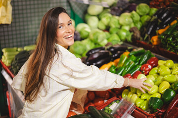 Young woman girl buyer client consumer stands in market store choosing food buying. Woman choose pepper, straight her hand. Customer choosing and buying fresh organic vegetables at food market.