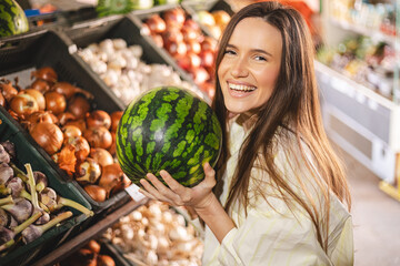Young woman girl buyer client consumer stands in shop store supermarket choosing food buying. Woman choose fruits, hold watermelon and look happy.