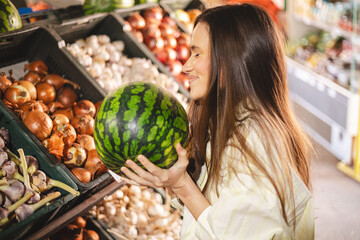 Young woman girl buyer client consumer stands in shop store supermarket choosing food buying. Woman choose fruits, hold watermelon and look happy.