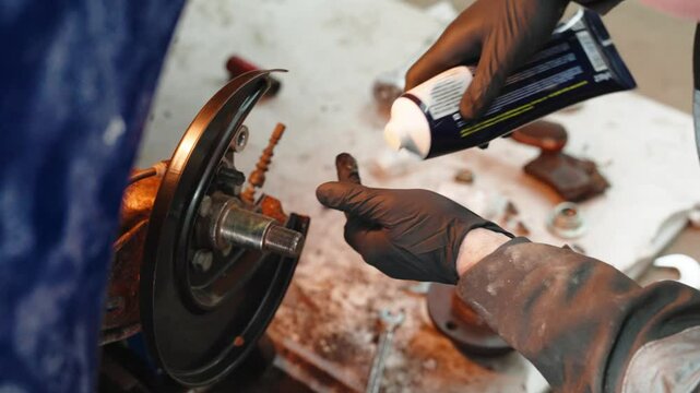 Skilled automotive technician wearing black protective gloves carefully applying white lubricating grease to car wheel hub, preventing friction and ensuring optimal vehicle performance