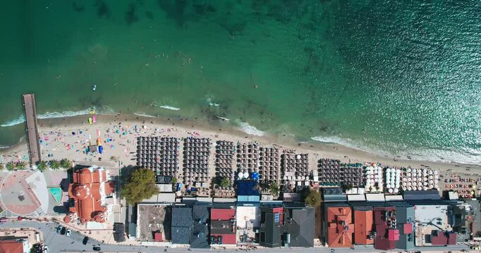 A high-angle aerial shot of Paralia, Greece, showcasing a bustling beach. Lined with umbrellas and overlooking vivid turquoise waters, tourists and locals alike enjoy this prime summer destination.