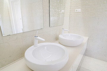 Bright bathroom featuring two modern, round white sinks, white faucets, light tile walls and mirrors.