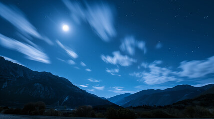 A bright full moon lights up soft clouds floating above towering mountains under a calm night sky.