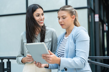 Two businesswomen standing outside of modern office building, using digital tablet working on new project discussing and explaining business matters and smiling. Design professional occupation persons