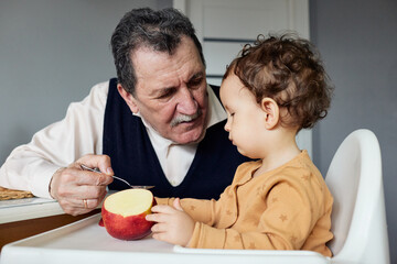 A grandfather feeds a juicy red apple slice to his curious grandson in their cozy kitchen on a sunny afternoon