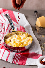 Scrambled Eggs with Cheese in a frying pan on a rustic table, a hand is sprinkling red pepper flakes over the eggs side view of simple breakfast dish