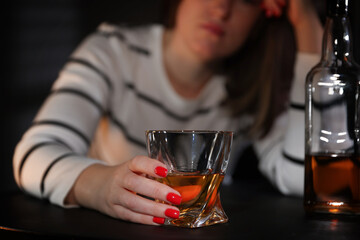 Alcohol addiction. Woman with glass of whiskey and bottle at table indoors, closeup