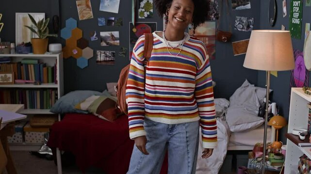 Tilt up slowmo portrait of cheerful African American teenage girl in colorful sweater with backpack standing in her room with maximalist interior expressing her individuality and smiling at camera