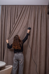 Woman ironing dark beige curtain with hand-held steamer