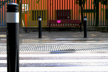 Paso de peatones a rayas blancas con pilonas y bolardos frente a un colegio con verja de colores y...