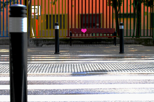 Paso de peatones a rayas blancas con pilonas y bolardos frente a un colegio con verja de colores y un banco de madera con coraz&oacute;n rosa pintado en una calle de Madrid