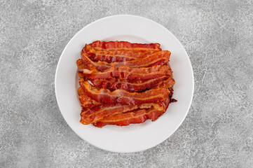Cooked bacon for breakfast on a white plate on a gray concrete background. Top view, copy space.
