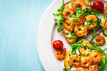 Toast with shrimp, arugula and avocado on a white plate with cherry tomatoes on a blue table. Space for text on a wooden background.