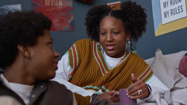 Rack focus of African American gen z boy engaged in conversation with his female friend sipping coffee from mug, sharing feelings and spending time together in cozy room at home