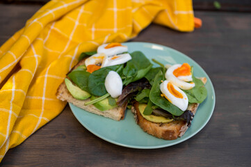 Delicious avocado toast with fresh spinach and soft-boiled eggs on rustic bread, served on a wooden table. A yellow checkered cloth adds warmth, making it perfect for healthy eating and brunch themes.