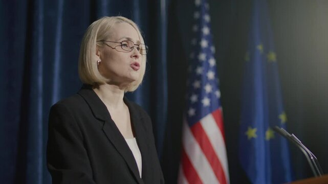 Middle-aged female diplomat delivering speech at podium with microphones during international summit with American and European Union flags in background