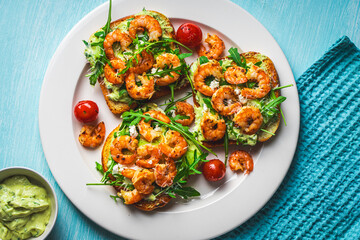 Toast or sandwiches with avocado oil, arugula and fried prawns on a white plate on a light blue background with a napkin. Healthy breakfast. View from above. Flat layout. Space for text.