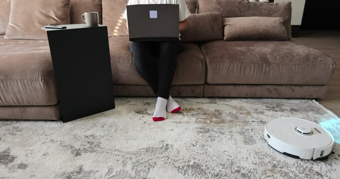 Young freelancer business woman using laptop, robot cleaner cleaning carpet. Detectors help to recognize walls and objects and optimize cleaning. Smart futuristic cleaning device. Technology concept