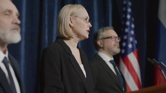 Female political leader standing at podium, accompanied by team, delivering speech to audience during press event with Russian and American flags on stage in background