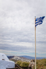 Vertical view from Pyrgos Kallistis, showcasing the Greek flag, cloudy sky, panoramic vistas of north Santorini, Aegean Sea, Greek islands, and coast, with whitewashed homes of Fira in the distance