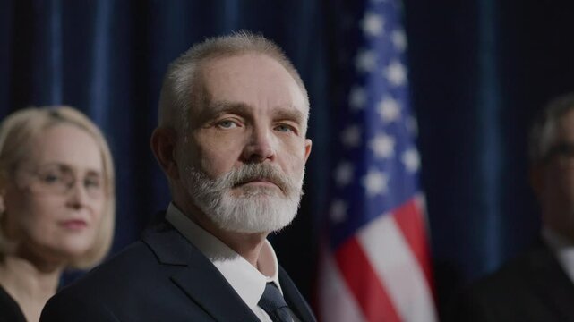 Senior male candidate in formal suit looking at camera with confidence during election campaign event with American flag and team members in background. Video portrait